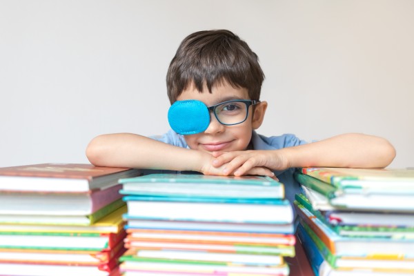 A child wearing a blue eye patch and glasses, leaning on a pile of books.