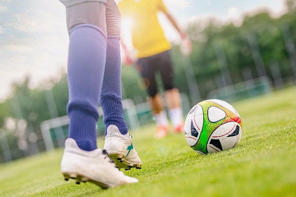 A close-up of a footballer wearing blue socks and white boots about to kick a colourful football on a grass pitch, with another player in the background in a yellow top.