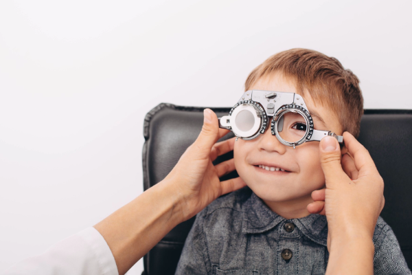 A smiling young boy wearing a trial frame during an eye test, with an optometrist's hands adjusting the frame.
