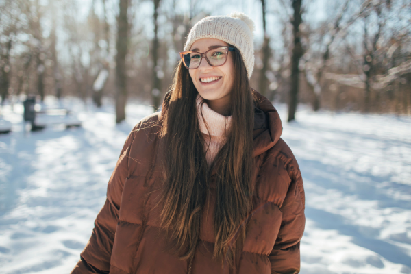 Smiling woman wearing glasses, a knitted hat and a brown puffer jacket, standing in a snowy wood on a sunny day